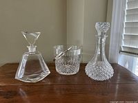 Three crystal bar set pieces on wooden surface: two decanters and an ice bucket with metal handle.