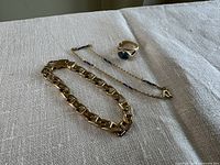 Thick yellow gold curb link bracelet, delicate chain bracelet, and gold ring with iridescent stone displayed on fabric.