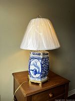 Full view of porcelain blue and white lamp with white pleated shade, lamp plugged in on wooden table against wall.