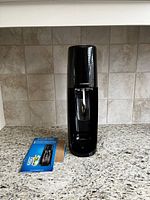 Front view of the black SodaStream sparkling water maker sitting on a granite countertop with the instruction manual beside it.