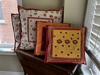 Two large cream floral print throw pillows with red piping in background, two smaller pillows in front with distinct warm tone floral covers