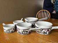 Five nesting porcelain pots with floral and fruit design, metal rims, and white handles arranged on a wooden table.