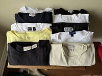 Top down view of 10 assorted women's cotton shirts stacked and folded, showing a range of colors mainly black, white, cream, and yellow.