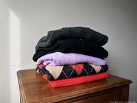 Stack of six women's sweaters folded on a wooden surface showing variety of colors and knit styles including argyle pattern and solid colors.