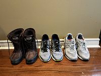 Photo of three pairs of women's shoes: two pairs of New Balance running shoes and one pair of UGG leather winter boots placed against a beige wall and wooden floor.