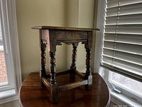 Front angled view of small wooden stool on round wooden table shows turned legs, rectangular top, and structural cross stretchers near base.