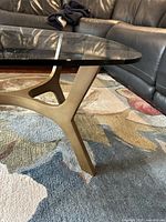 Close-up side view of brass-finish sculptural cast aluminum base supporting clear glass top on carpet.