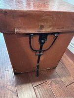 Elevated side view showing metal latch and handle attached to reddish-brown box with riveted corners.