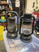 Two vintage railroad lanterns on a plastic container in a garage, one clearly larger and bulkier with black paint, the other smaller and slimmer with visible rust.
