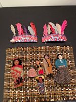 Seven dolls, woven placemat, and two vintage feathered headresses on a black surface