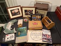 Overview of lot items arranged on table including books, framed fish art, fish-shaped thermometer, cooler, wooden spool, cigar box, and carved fish figurines
