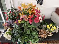 Various mixed artificial and dried flowers arranged on a table, showing different colors and types including lilies, daisies, begonias and green leaves.