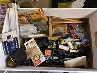 Top-down view of box with assorted garage items including packaged fishing lures, hand tools with wooden handles, and other miscellaneous objects.