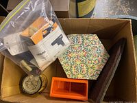 Box containing the glue tool packaging, a hexagonal tile with floral pattern, a sealed canister and small parts in a plastic bag.