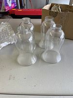 Four clear glass hurricane lampshades arranged on a table with a cardboard box in background.