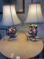 Pair of vintage porcelain table lamps with detailed figurines of man and woman, each with fabric lampshades trimmed with lace, placed on round table.