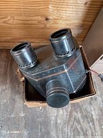 Front view of the Radioptican postcard projector showing the metal body with two large cylindrical lenses on top and one lens at front center, placed inside a cardboard box.