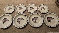 Overview of 8 stoneware plates with rainbow trout design on carpet.