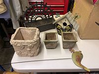 All items displayed on a white table including large and small stone planters, decorative birdhouse with butterfly decorations, and bird figurine.
