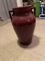 Maroon ceramic floor vase placed on carpeted floor in front of grey curtains, shows full front and one handle.