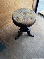 Photo showing top and side of the round padded seat with floral fabric upholstery and button tufting, on wooden tripod base with carved legs.