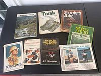 Photo of seven vintage outdoor books laid out on a black surface, titles visible including hunting, fishing, wilderness and military tank themes