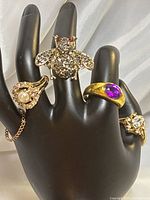 Rings displayed on a black mannequin hand showing the bee-shaped ring, pearl ring, purple stone ring, and another rhinestone ring.
