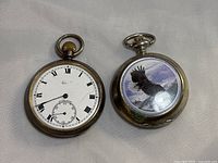 Two silver pocket watches side by side on fabric background; one shows white dial with Roman numerals, the other shows decorative eagle on cover.