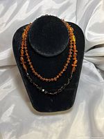 Two amber beaded necklaces displayed on black bust stand on satin fabric background. Orange amber necklace approx. 12 inches, and orange/cherry amber necklace approx. 14 inches.