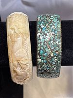 Front view of two bangle bracelets on black display stand showing detailed bone carving and turquoise mosaic inlay.