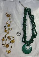 Two necklaces laid flat on white fabric background with a quarter for size reference; one is a green malachite beaded 3-strand necklace with a round pendant, the other is a gold-tone chain with assorted oval and round textured beads.