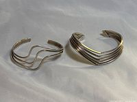 Two sterling silver cuff bracelets side by side on white cloth background, showing the V-shaped and wavy designs