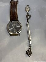 Two watches side by side on a light background: Kenneth Cole with black dial and brown leather strap; Timex smaller with silver tone face and sterling silver band showing floral decorations.