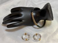 Leather cuff bracelet displayed on a black hand stand with two-tone copper and silver details and wire wrap decoration. Pair of two-tone metal hoop earrings with gold accents placed beneath the stand.