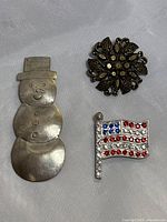 Set of three brooches including a silver-tone snowman, dark metal flower, and rhinestone American flag laid on white fabric.