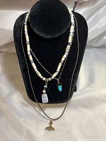 Three necklaces displayed on a black necklace stand: blue and white agate beaded necklace with white agate pendant, sterling chain with turquoise and onyx arrowhead pendant, brown leather cord with bronze pendant.
