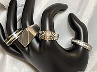 Four men's rings displayed on black hand stand, showing various styles and details.