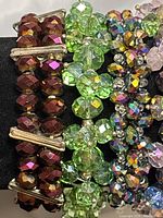 Close-up photo of five beaded bracelets showing colors and bead details: dark reddish-purple with gold spacers, light green, multicolor iridescent, pale purple, and mixed-color beads with turquoise and shell accents.