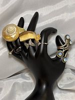 Front and side angle view of all 7 rings displayed on black hand-shaped stand, showing varied designs and colors including conch, flower, silver, black, wood and bone, and opaline styles.