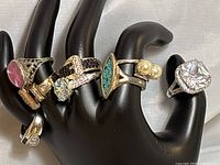Front view of 6 rings on a black hand display, showing various stones and metallic finishes including clear, turquoise, pearl, black stones, and pink cabochon.