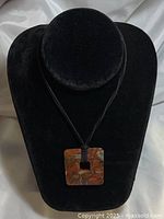 Black leather cord necklace with square polished agate or jasper pendant with a central cut-out displayed on black velvet necklace stand.