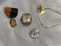 Photo of four items on white cloth: Hamilton watch case, sports medal on ribbon, gold tone necklace with pendant, and a quarter coin for scale.