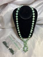 Jade necklace displayed on a black stand, showing round light green beads and decorative pendant. Small clear plastic bag of loose dark green beads nearby.