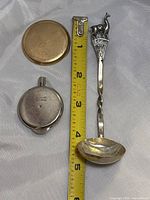 Photo showing the gold round watch back, round map measuring device marked Tacpo Inc. Tillamook, and silver souvenir spoon with twisted handle and llama figure on top labeled 'PERU'