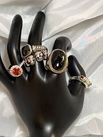 Five silver plated rings displayed on black hand mannequin, showing variety of designs including colored and clear stones, and skull motifs.