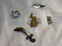 Five brooches arranged on white fabric: silver sword, mixed metal fairy, circular rhinestone, leaf with hematite, butterfly brooch