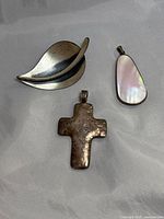 Three jewelry pieces displayed on fabric: leaf-shaped silver brooch, hammered cross pendant, and teardrop-shaped abalone pendant with silver frame.