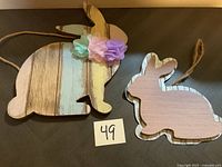 Two wooden bunny decorations displayed side by side on a flat surface with label '49'. Larger bunny has pastel vertical stripes and fabric flowers; smaller bunny is solid pastel pink with natural wood edges.