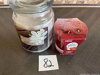 Large glass jar candle labeled 'Paradise Vanilla' next to unopened Glade plug-in air freshener box with warm apple pie scent indicated.