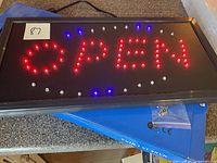 LED OPEN sign illuminated showing red OPEN letters amid blue and white lights, laying flat on a surface with power cord visible.